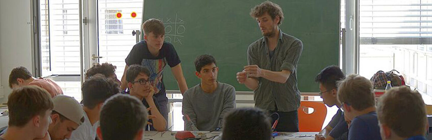 a group of students is gathered around a scientist who concentrates on something in his hands